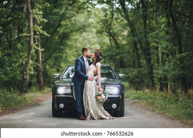 Newlyweds Near The Black Wedding Car Stand On The Road In The Summer Forest.