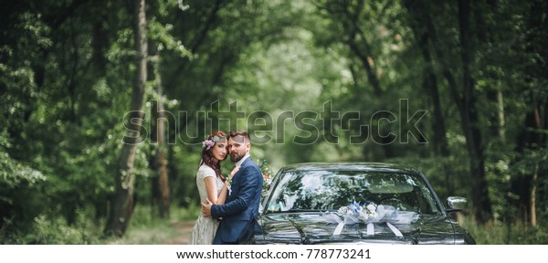Newlyweds near the black wedding\
car stand on the road in the summer forest. Panorama, copy\
space.