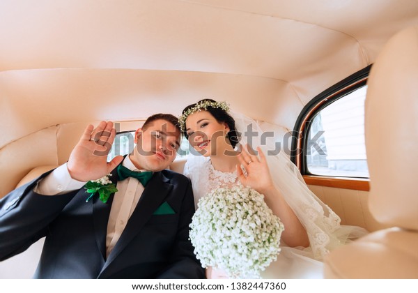newlyweds are sitting in the car waving their\
hands and looking at the camera. car interior. Bride holding\
wedding bouquet