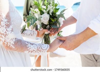 Newlyweds At The Wedding Romantic Couple Holding Hands During Destination Wedding Marriage Matrimonial Ceremony On The Sandy Beach In Dominican Republic, Punta Cana. Family, Love, Unity Concept.  