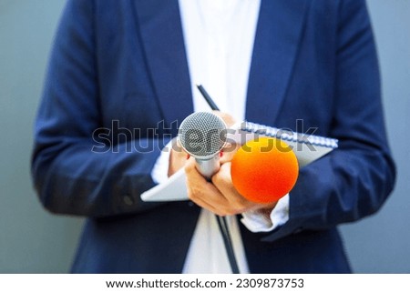 News reporter at media event or press conference, holding microphone, writing notes. Broadcast journalism concept.