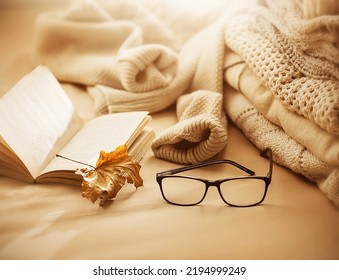 Next To A Stack Of Warm White Wool Sweaters Is An Open Interesting Book, Glasses And A Red Dry Maple Leaf, Illuminated By The Rays Of The Sun. Home Comfort And Relaxation In The Cold Days Of Autumn.