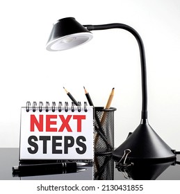 NEXT STEPS Text On Notebook With Pen And Table Lamp On Black Background