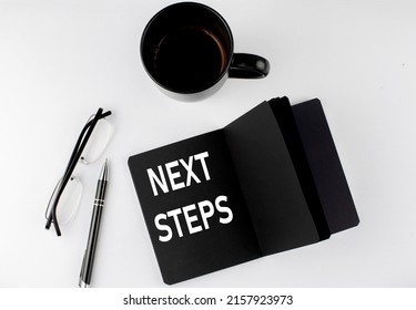 NEXT STEPS Written Text In Small Black Notebook With Coffee , Pen And Glasess On A White Background. Black-white Style