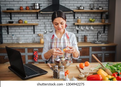 Nice adult woman sit at table in kitchen. She break eggs and pour them into bowl. Model look down. She concentrated. Woman wear apron.