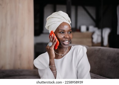 Nice African girl in turban talks by phone home smiles wide happy to hear friend, missing them after relocation. Successful African American businesswoman in traditional clothes speaks by smartphone.