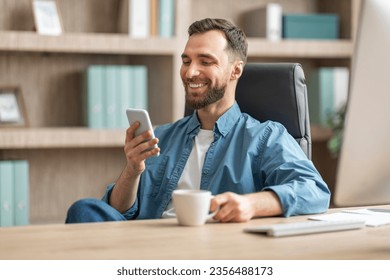 Nice App. Young Businessman Relaxing With Smartphone And Coffee At Workplace, Cheerful Millennial Male In Casual Clothes Sitting At Desk, Holding Mug With Hot Drink And Messaging On Mobile Phone - Powered by Shutterstock