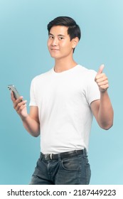 Nice Asian Guy In White T-shirt And Jeans Holding Smartphone And Show Thumbs Up Isolated On Blue Background