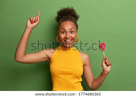 Nice attractive carefree girlfriend dances happily with heart shaped lollipop, has fun indoor, has sweet tooth and good mood after eating yummy candy, moves with joy over vivid green background