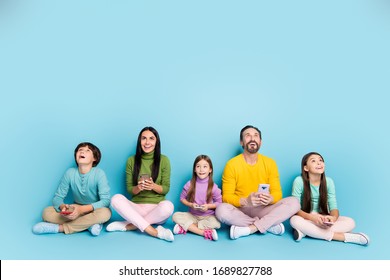 Nice attractive lovely charming cheerful cheery big full family pre-teen kids sitting in lotus pose using gadget looking up copy space isolated on bright vivid shine vibrant blue color background