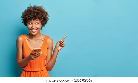 Nice attractive young Afro American woman uses cell phone and points on copy space shows direction dressed in stylish orange dress isolated over blue background. Ethnic lady with modern device