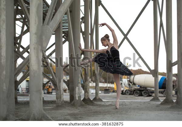 Nice\
ballerina posing leaning on a concrete support structure. She\
stands on the toe of the left foot in black tutu. Behind her is a\
parking for technical cars. Outdoor.\
Horizontal.