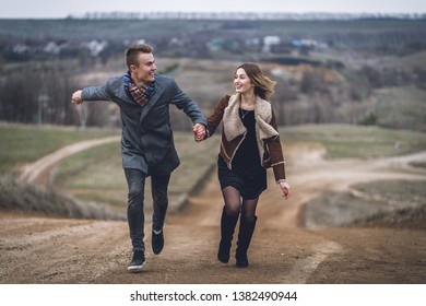 A Nice Beautiful Lovely Couple Running In The Park Or Field. Man In Gray Coat And Scarf With Girl Woman In Brown Coat. L. Lovers Run Away Holding Hands.