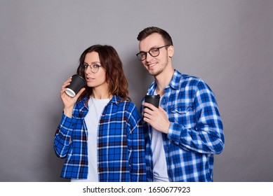 A Nice Beautiful Young Caucasian Couple, He, She, In Blue Plaid Shirts And Glasses, Squiggle Black Coffee Cups, Look Into The Camera And Smile.