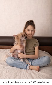 Nice caucasian girl in a green t-shirt is sitting on a bed with her adorable hairless dog pet. Chinese crested puppy, family indoor activity, portrait. Front view.