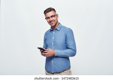 A nice chat. Portrait of handsome and cheerful bearded man in casual wear holding smart phone and looking at camera while standing against grey background