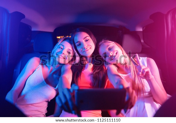 Nice and cheerful girl are sitting in car\
together. Blonde girls are resting on their friend\'s shoulders.\
Brunette is holding black phone. She is taking selfie. Young women\
are looking on phone.
