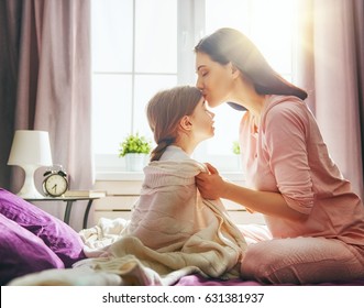 A Nice Child Girl And Her Mother Enjoy Sunny Morning. Good Time At Home. The Mom Wakes The Kid With A Kiss On The Bed In The Bedroom.