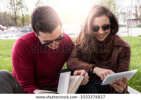 nice couple boyfriends relaxing sitting on meadow looking at book and smart phone