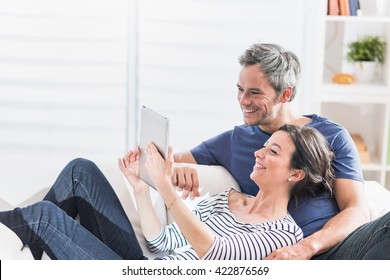 a nice couple at home using a tablet, they are sitting on a white couch, having fun by sharing a video on the web