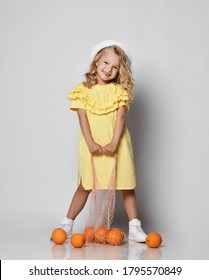 Nice Cute Smiling Blonde Curly Kid Girl In Yellow Dress, White Sneakers And Hat Stands Holding Bag With Oranges On The Floor. Stylish Comfortable Casual Fashion For Children Concept