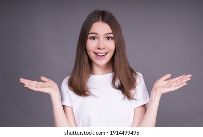 Nice cute young Caucasian girl with dark hair, surprised, spread her arms to the sides, opened her mouth and looks straight, wears a bob haircut and a white T-shirt, photo on a gray background. - Shutterstock ID 1914499495