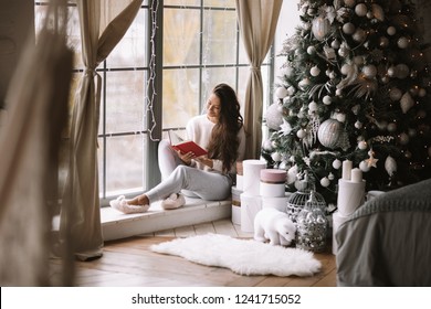 Nice dark-haired girl dressed in pants, sweater and warm slippers reads a book sitting on the windowsill of a panoramic window in the room next to the New Year tree, gifts and candles