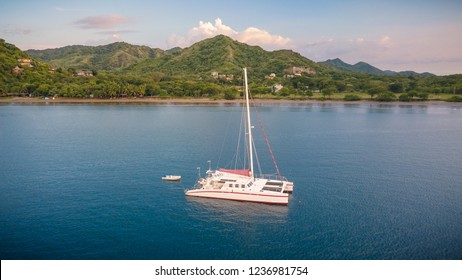 A nice drone shot of a large white catamaran yacht anchoring in a beautiful blue bay in front of an empty beach on the green coast of Costa Rica in Central America.