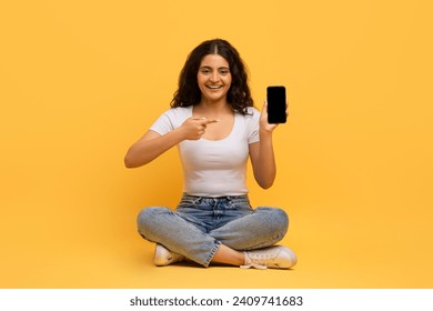Nice educational mobile application, online studying. Cheerful attractive curly young hindu lady sitting on floor with smartphone in her hand, yellow studio background