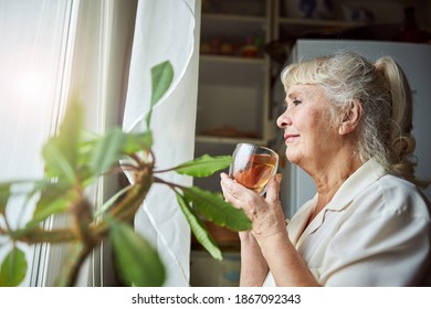 Nice elderly lady looking out the window and smiling while holding cup of hot drink - Powered by Shutterstock