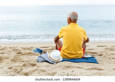 Nice elderly man resting and looking to something interesting. Clothes on the sand near the sea