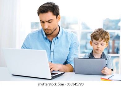Nice family photo of little boy and his father. Boy and dad sitting at room with big window. Young man working with laptop while boy using tablet computer