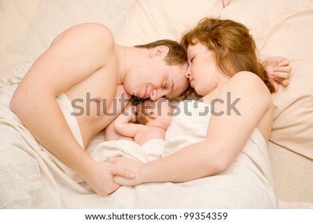 Nice family sleeping together in a white bed. Mother, father and newborn baby. Baby eating the mother's breast. Happy dream. Woman and man holding hands
