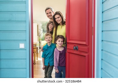 A Nice Four People Family Is Opening Their Stylish Red Door To Welcome The Guest. The Parents And Their Two Children Are Smiling And Wearing Casual Clothes. The House Have Blue Wall And Wooden Floors