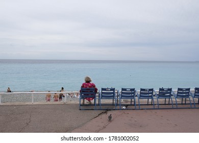 Nice, France - 06 18 2020: An Old Lady Sitting From Behind On The Famous Blue Chairs Of The Promenade Des Anglais Looks At The Beach And The Sea In June 2020