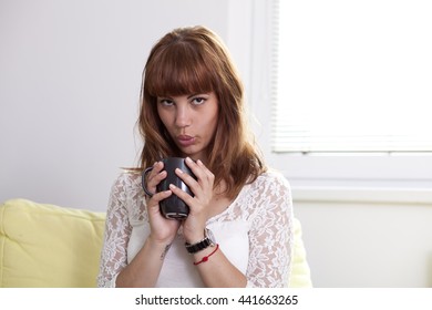 Nice girl drinking a cup of hot coffee or tea and blowing at it, look straight at you, brunette with white dress on yellow couch near window with sun on hair casting rim light