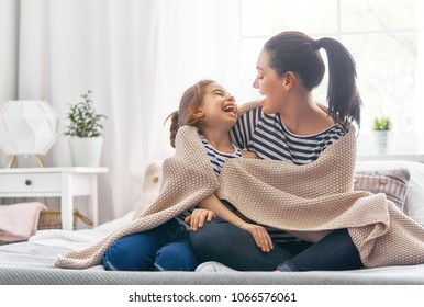 A nice girl and her mother enjoy sunny morning. Good time at home. Child wakes up from sleep. Family playing under blanket on the bed in the bedroom.