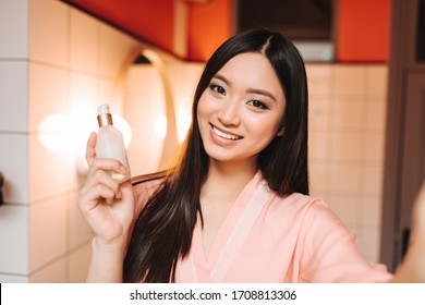 Nice girl holds cosmetics for face. Lady smiles and takes selfie in bathroom - Powered by Shutterstock