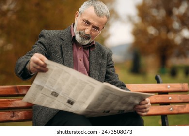 A nice grandfather with a beautiful beard in a gray jacket sits on a bench in the park and reads a newspaper - Powered by Shutterstock