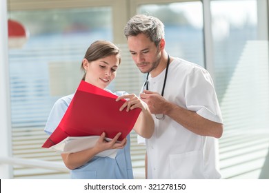 A Nice Grey Hair Doctor With Beard And A Nurse Are Examining A Patient File He Is Wearing His White Coat, His Stethoscope Around His Neck. They Are Standing In Front Of Hospital Room With Glass Walls