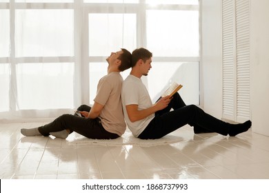 A Nice Guy Reads A Book And His Friend Listens To Him And Looks Into A Smartphone. Gay Couple Of Men Spend Time At Home Sitting On The Floor Of The House In A Light Interior