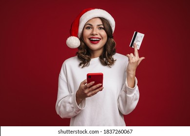 Nice happy girl in Santa Claus hat posing with credit card and mobile phone isolated over red background - Powered by Shutterstock