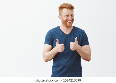Nice job man, like it. Pleased good-looking happy male friend with ginger hair and brislte, showing thumbs up and smiling broadly, giving positive feedback, sharing his positive opinion over grey wall