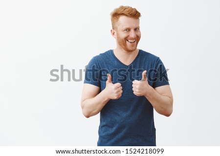 Nice job man, like it. Pleased good-looking happy male friend with ginger hair and brislte, showing thumbs up and smiling broadly, giving positive feedback, sharing his positive opinion over grey wall