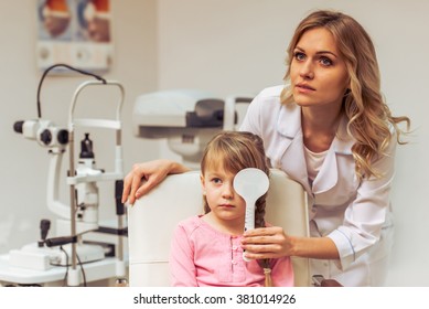 Nice Little Girl At The Ophthalmologist On Consultation. Beautiful Female Doctor Covering Girl's Eye And Checking Her Visual Acuity