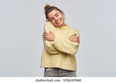 Nice looking woman, beautiful girl with blond hair gathered in bun. Wearing yellow sweater. Hugs herself, feel warm and comfortable. Keeps eyes closed. Stand isolated over white background