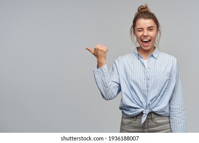 Nice Looking Woman, Beautiful Girl With Blond Hair Gathered In Bun. Wearing Striped Knotted Shirt. Pointing With Thumb To The Left At Copy Space And Wink To The Camera, Isolated Over Grey Background