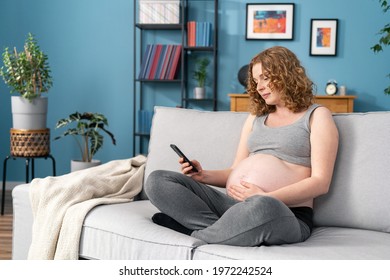 Nice Lovely Sweet Tender Beautiful Cheerful Pregnant Curly-haired Mom Sitting On Sofa, Couch, Holding Belly, Working Remotely On Cell Phone, Lotus Pose