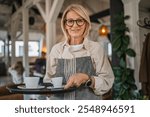 Nice mature woman waitress in apron offer cup of delicious tasty coffee, stand and smile, friendly staff of restaurant