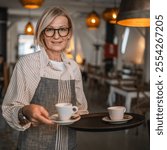 Nice mature woman waitress in apron offer cup of delicious tasty coffee, stand and smile, friendly staff of restaurant